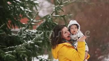 可爱的年轻女子穿着黄色<strong>羽绒服</strong>和婴儿穿着工作服走路。 下雪了，妈妈给他的儿子看，